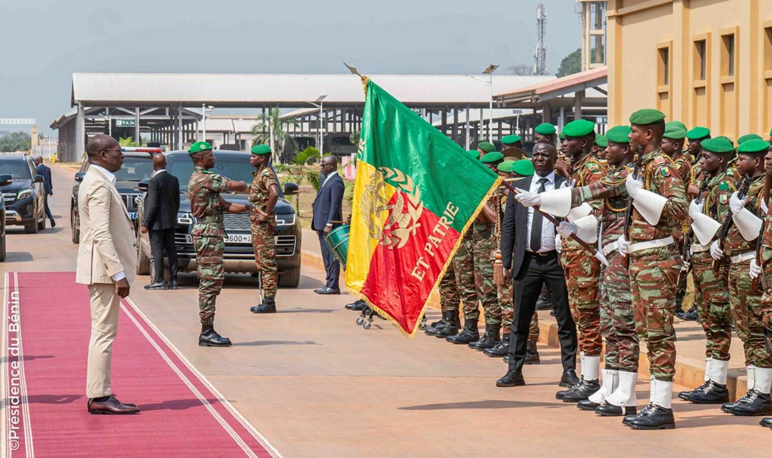 Un homme en costume clair salue une formation de soldats en tenue camouflage et bérets verts avec un drapeau tricolore sur un tapis rouge extérieur