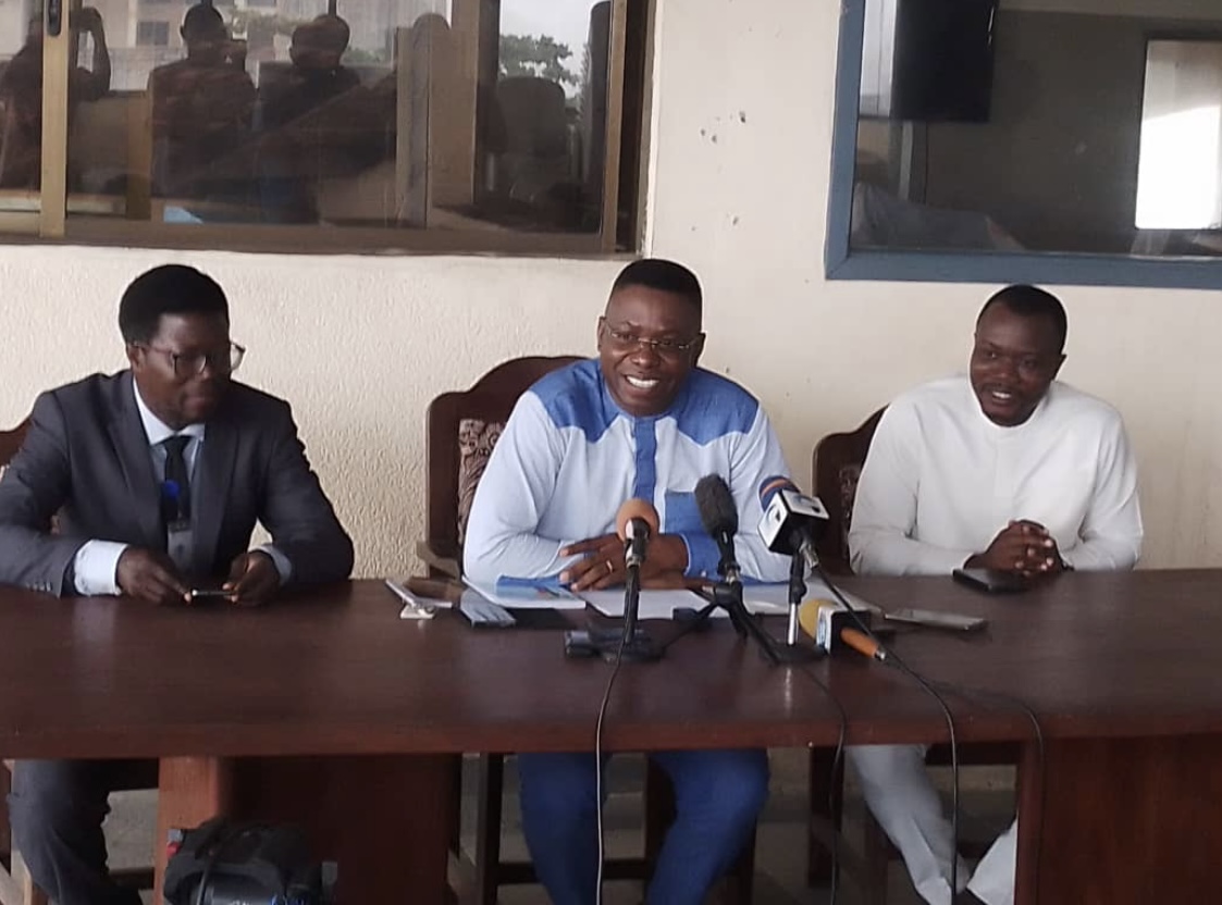 Trois hommes assis à une table devant des micros, discutant lors d'une conférence ou d'une réunion