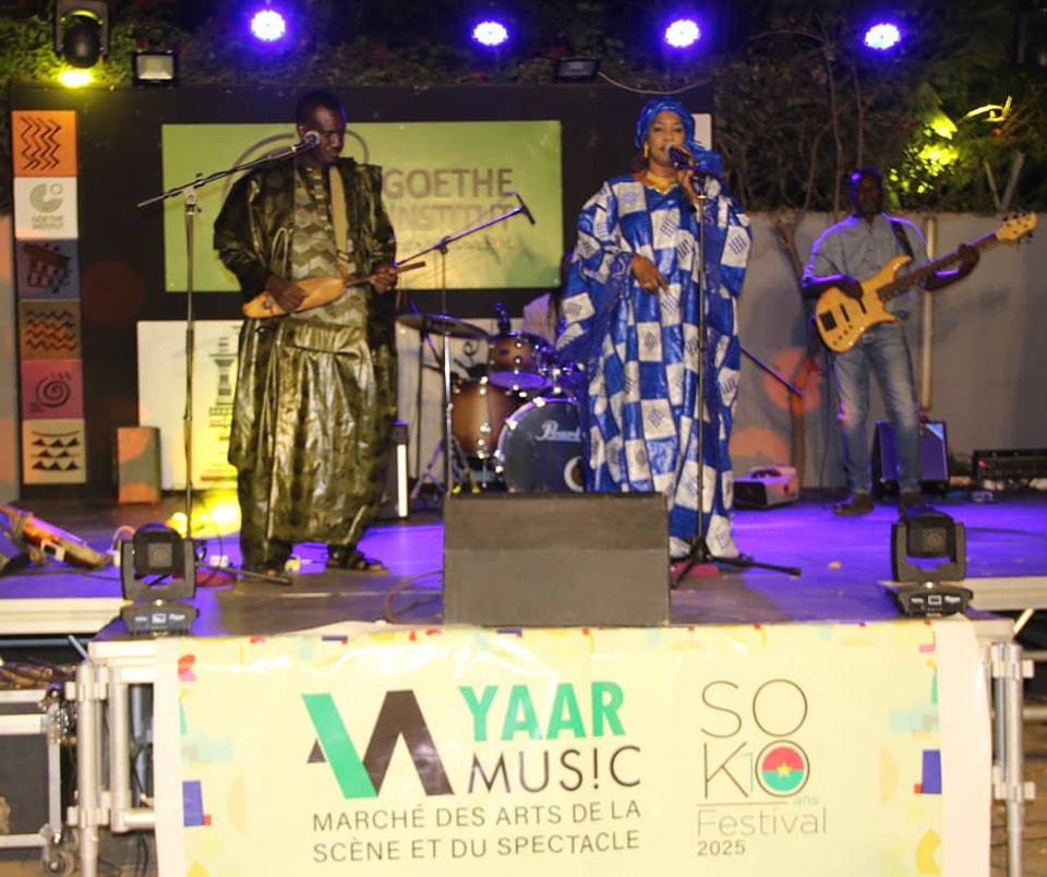 Trois musiciens en tenue traditionnelle jouent et chantent sur scène lors d’un festival de musique en soirée