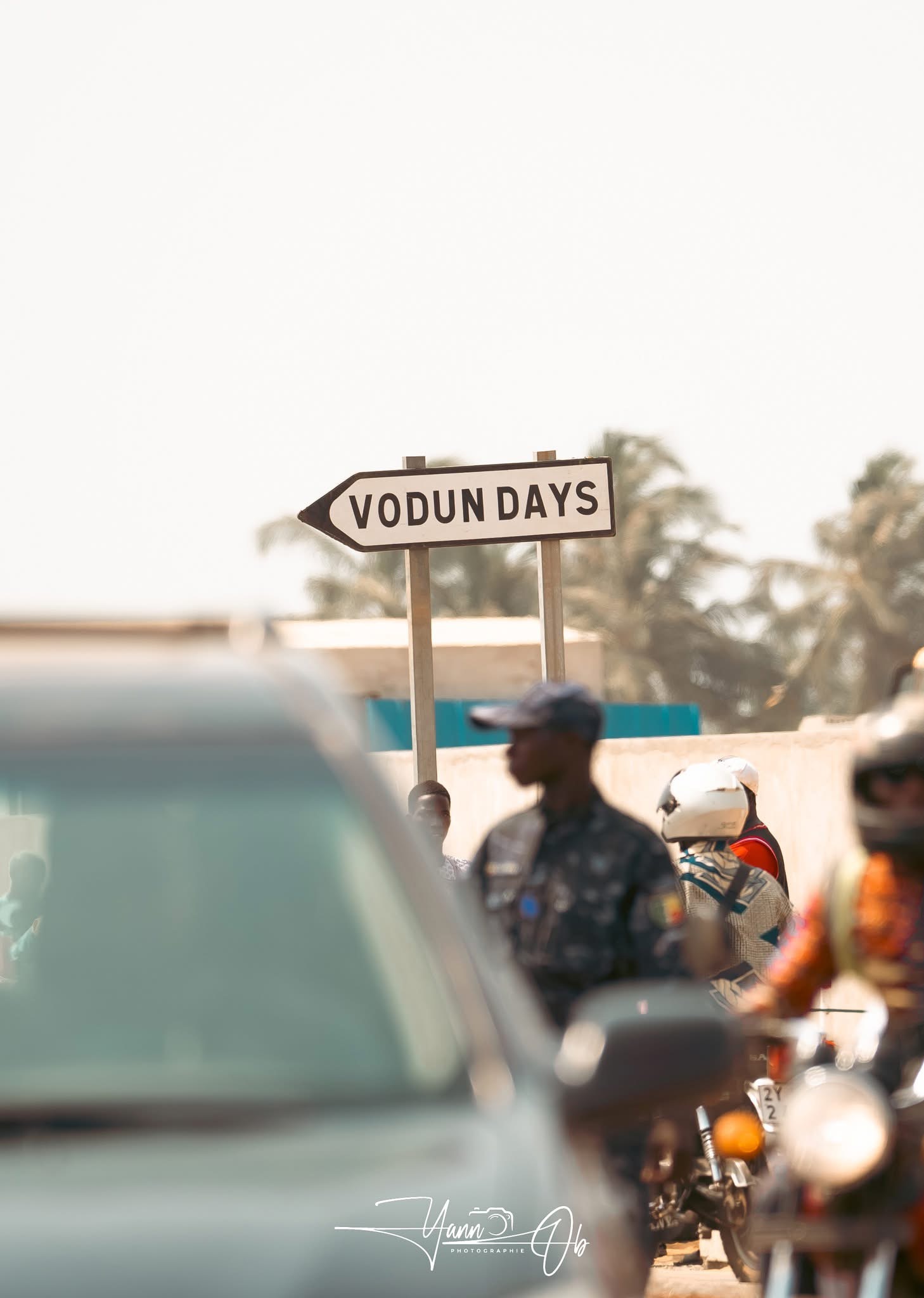 Panneau directionnel « VODUN DAYS » tenu au-dessus d'un trottoir avec des passants et des véhicules flous au premier plan