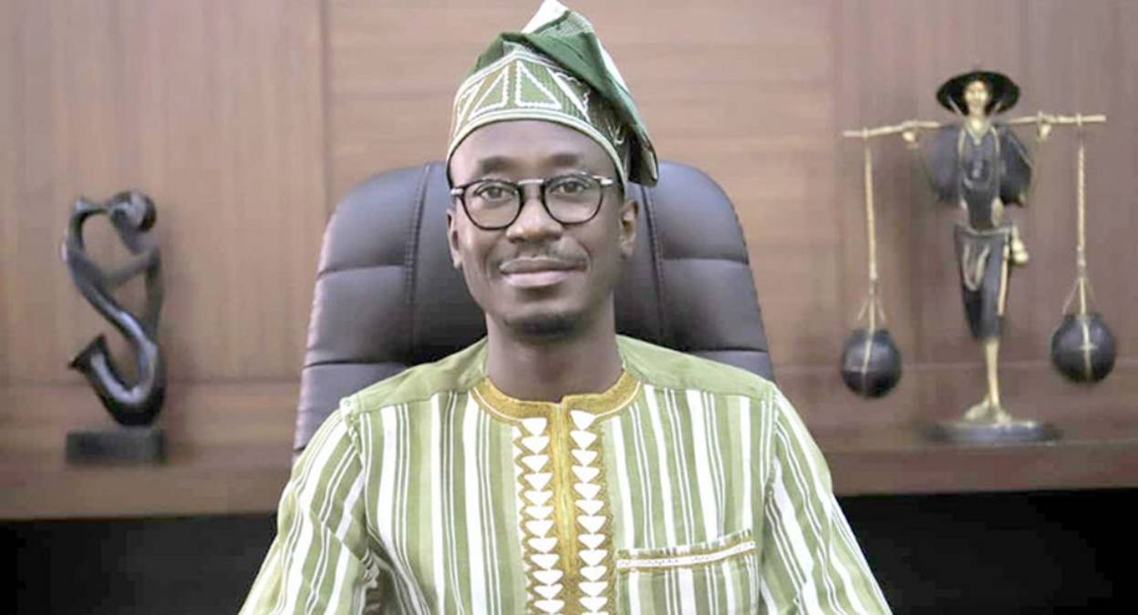 Homme souriant en tenue traditionnelle rayée et chapeau assis devant un bureau avec deux sculptures sur une étagère en arrière-plan
