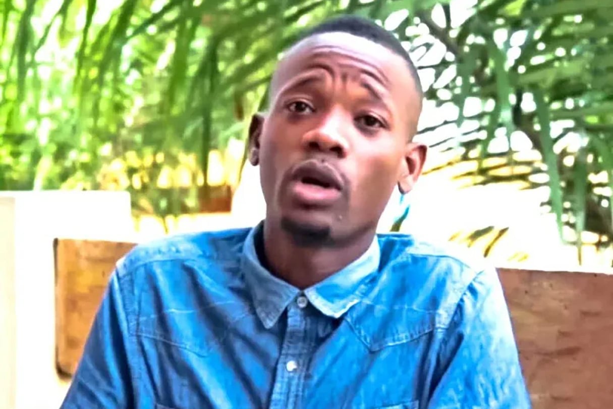 Jeune homme en chemise bleue parlant, assis devant un fond de feuillages verts et lumière naturelle