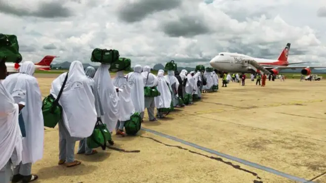 Une file de personnes habillées en blanc avec sacs verts fait la queue sur un tarmac près d’un avion au ciel nuageux