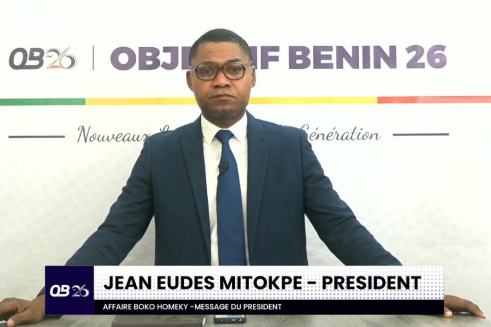 Un homme en costume bleu et cravate se tient devant un fond avec le texte OB26 OBJETIF BENIN 26