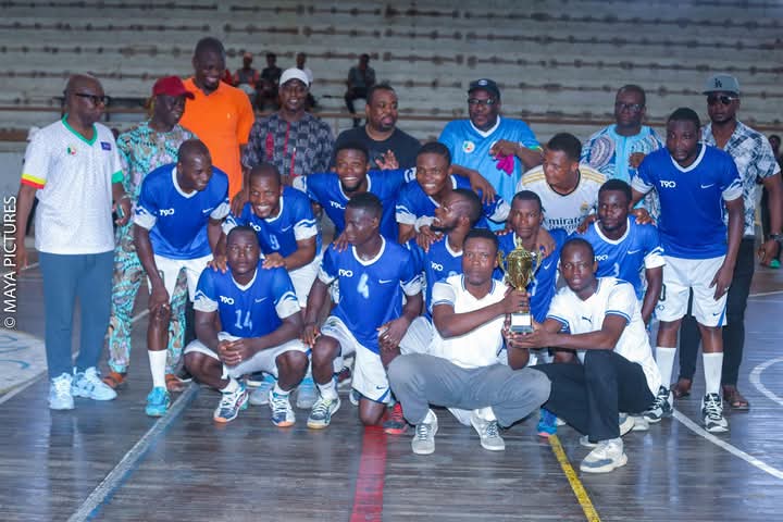 Équipe de joueurs en tenue bleue posant avec un trophée au milieu d’un terrain couvert avec des personnes debout derrière eux