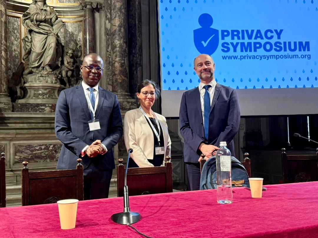 Trois personnes debout derrière une table rouge avec micros et gobelets dans une salle ornée et un écran Privacy Symposium en arrière-plan