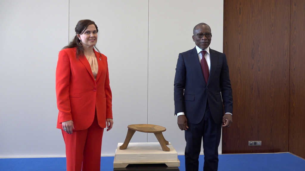 Un homme en costume sombre et une femme en tailleur rouge posent debout près d’un petit tabouret en bois sur un socle beige