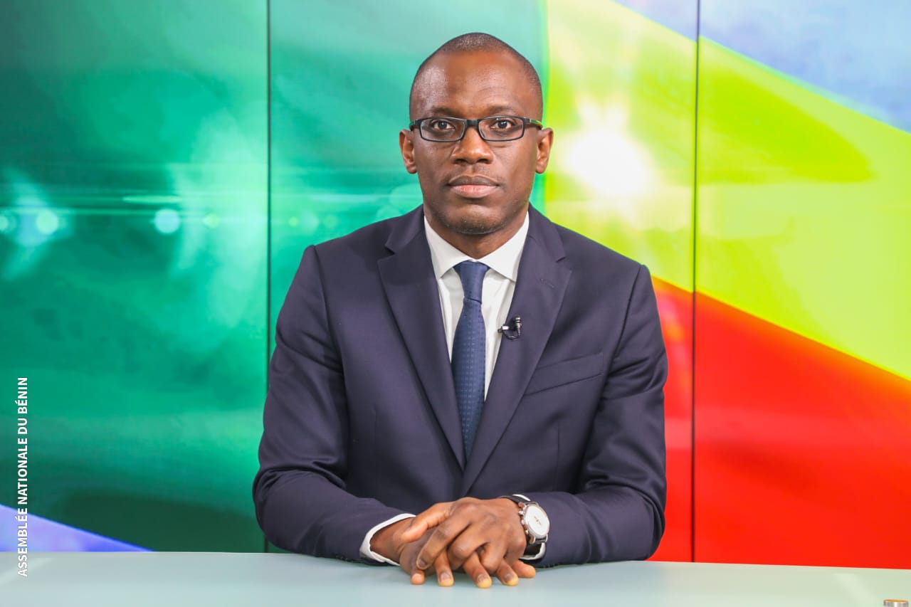 Homme en costume sombre et lunettes assis devant un bureau avec un fond coloré vert et jaune rouge