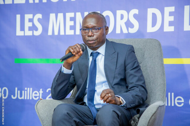 Homme en costume bleu parlant au micro assis sur un fauteuil devant un fond bleu avec du texte blanc