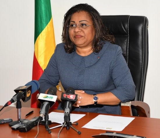 Une femme assise derrière un bureau avec plusieurs microphones et un drapeau aux couleurs vert, jaune et rouge en arrière-plan