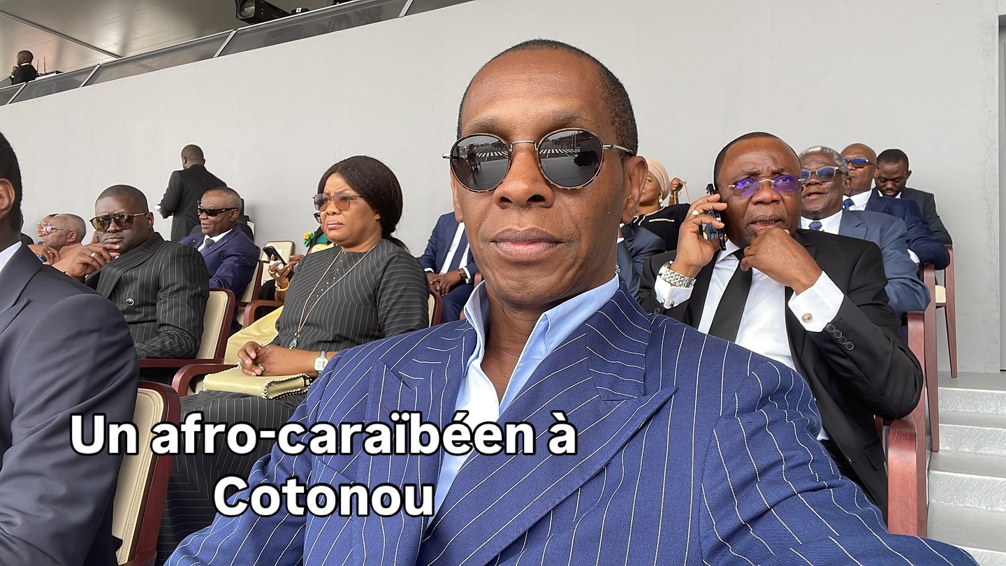 Un homme en veste rayée bleue et lunettes noires assis dans une tribune avec plusieurs autres personnes derrière lui