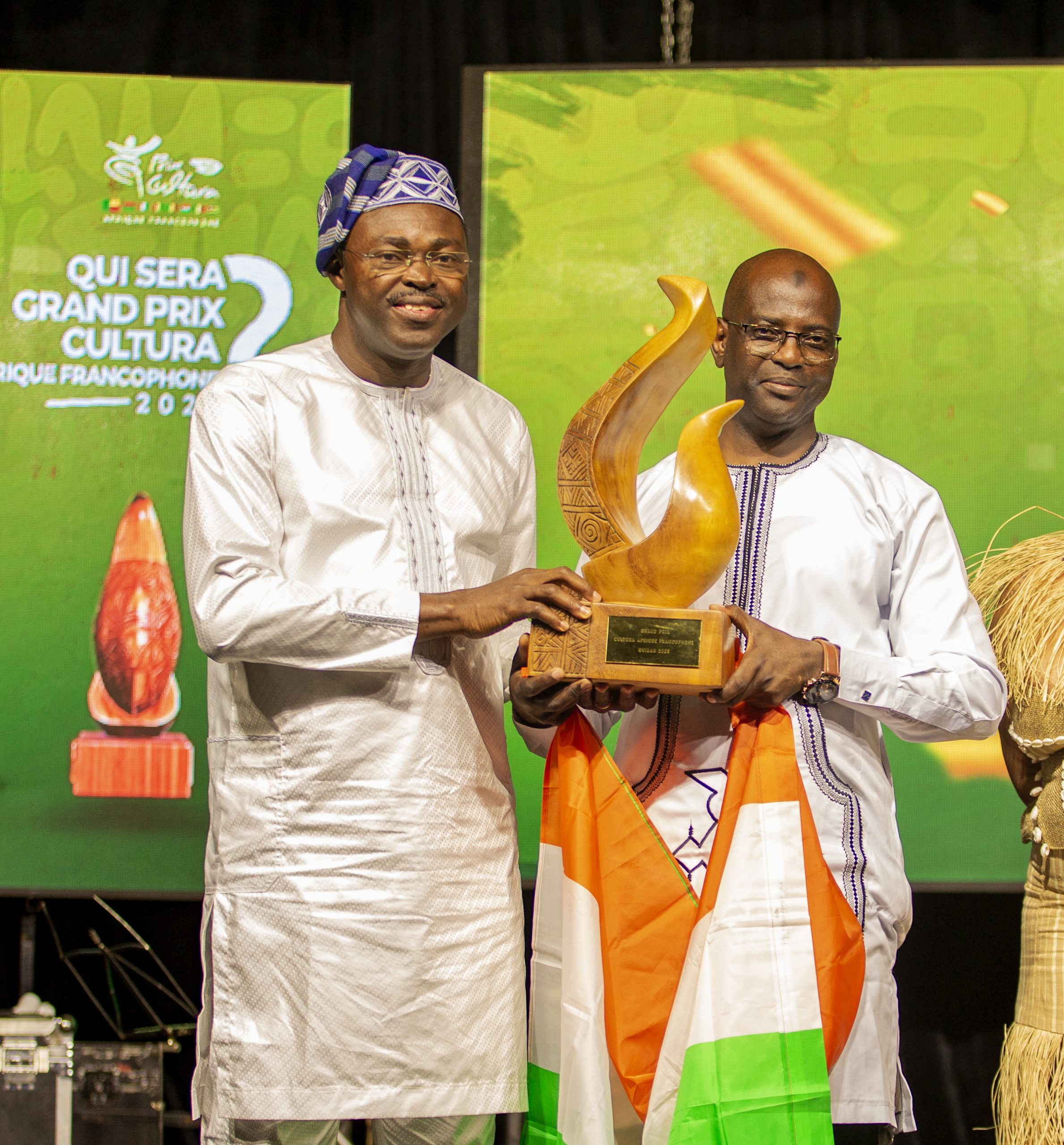 Deux hommes en tenue traditionnelle tiennent ensemble un trophée en bois sur une scène avec un drapeau orange blanc vert posé sur un bras