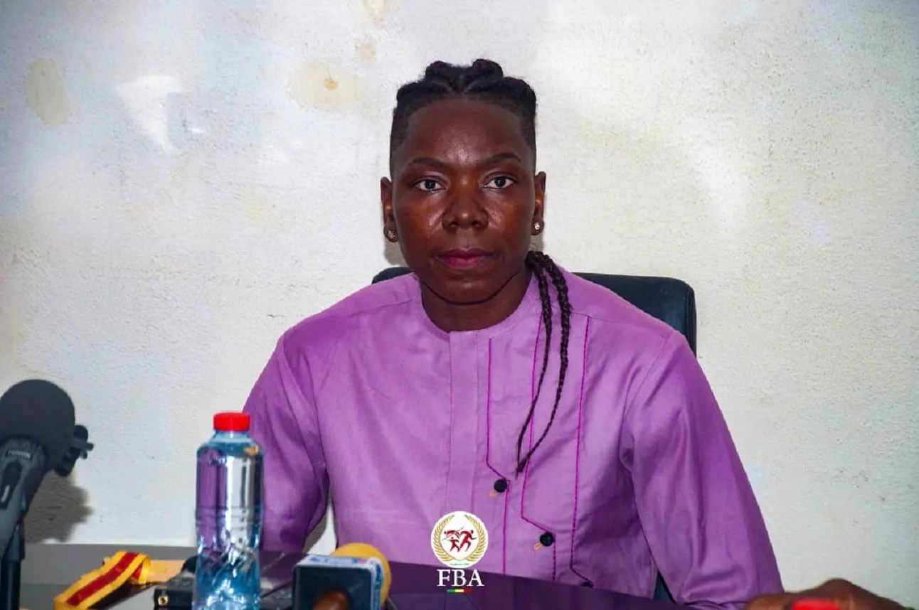 Femme assise à une table devant un mur clair, vêtue d’une tunique violette avec plusieurs microphones devant elle
