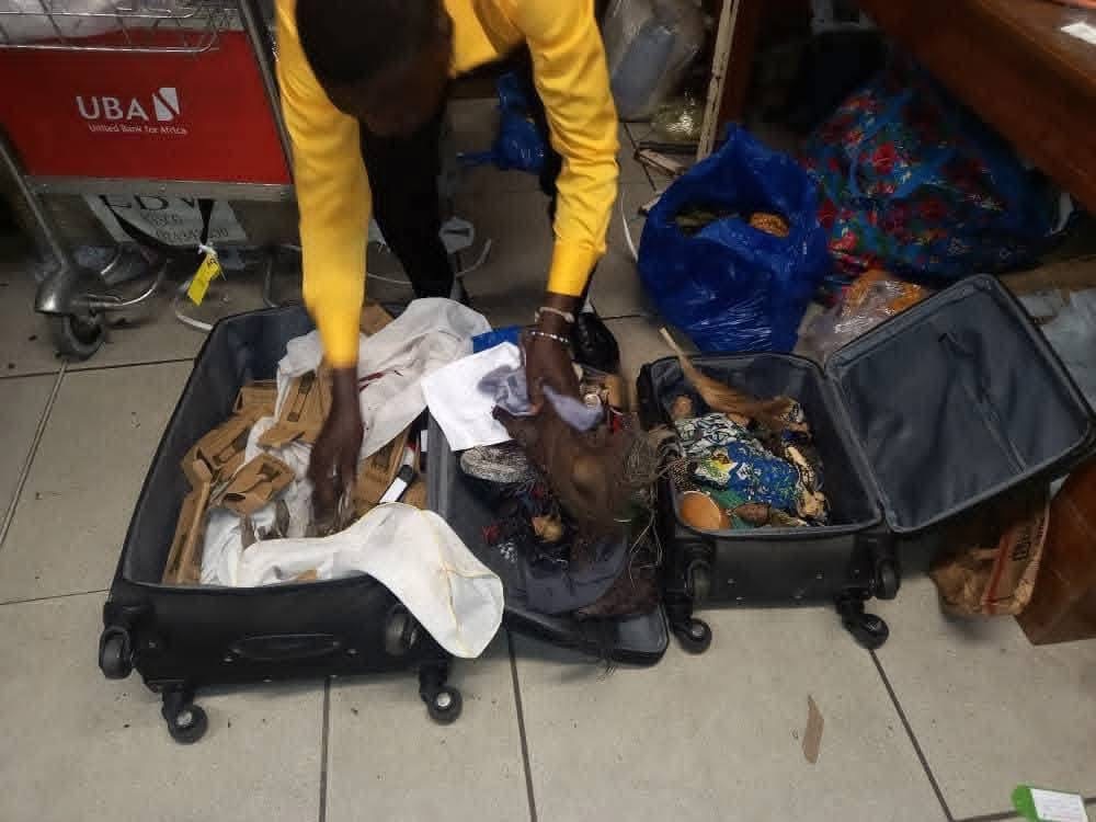 Personne en chemise jaune manipulant deux valises ouvertes contenant divers objets artisanaux et tissus colorés sur un sol carrelé