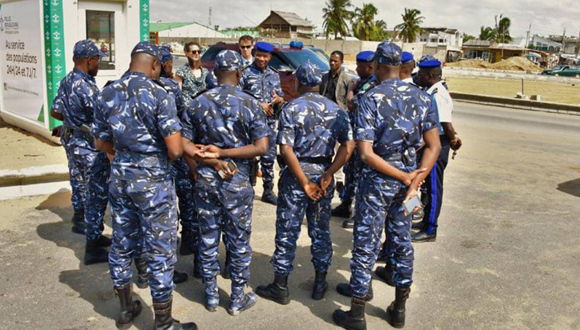 Un groupe de policiers en uniforme bleu camouflé est rassemblé en cercle sur une chaussée face à plusieurs passants.