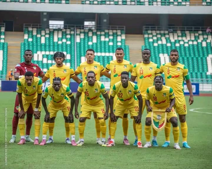 Onze joueurs de football en tenue jaune posent sur un terrain dans un stade avec des sièges verts et blancs vides