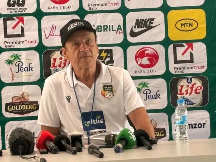 Homme en polo blanc et casquette noire assis devant des micros et un mur de logos publicitaires verts