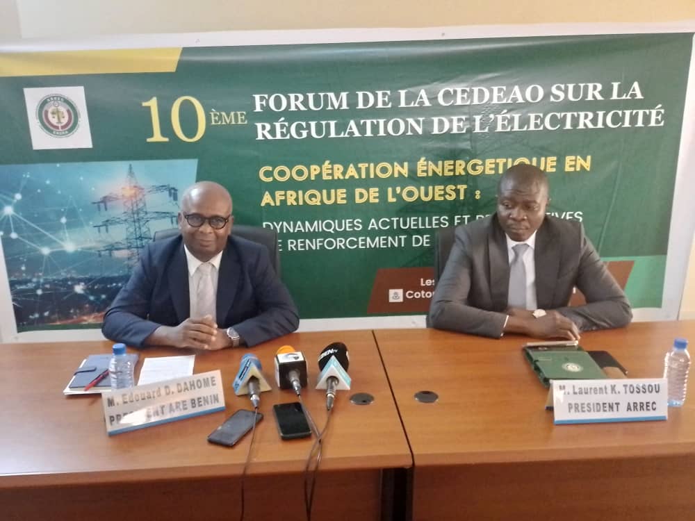Deux hommes en costume assis derrière une table avec des microphones et une banderole de forum sur la régulation de l’électricité en arrière-plan