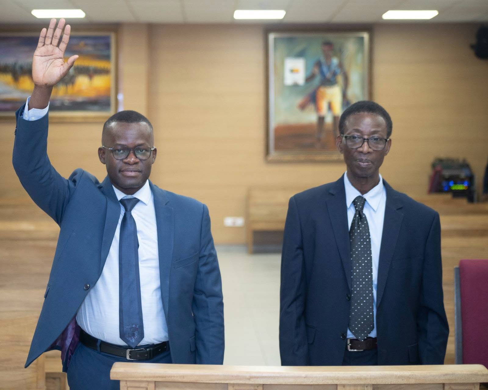 Deux hommes en costume debout dans une salle, l’un lève la main droite
