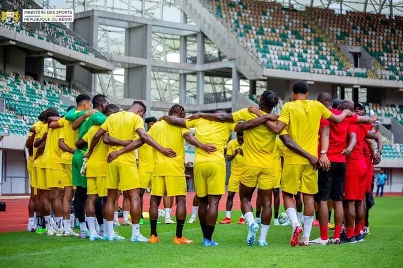Joueurs en tenue sportive jaune en cercle sur un terrain de football dans un stade aux gradins vides partiellement occupés