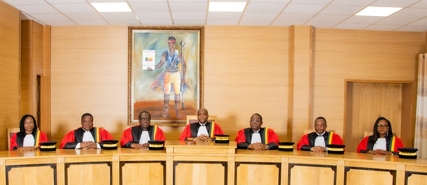 Sept personnes en robe officielle sont assises derrière une grande table en bois avec un tableau accroché au mur derrière eux