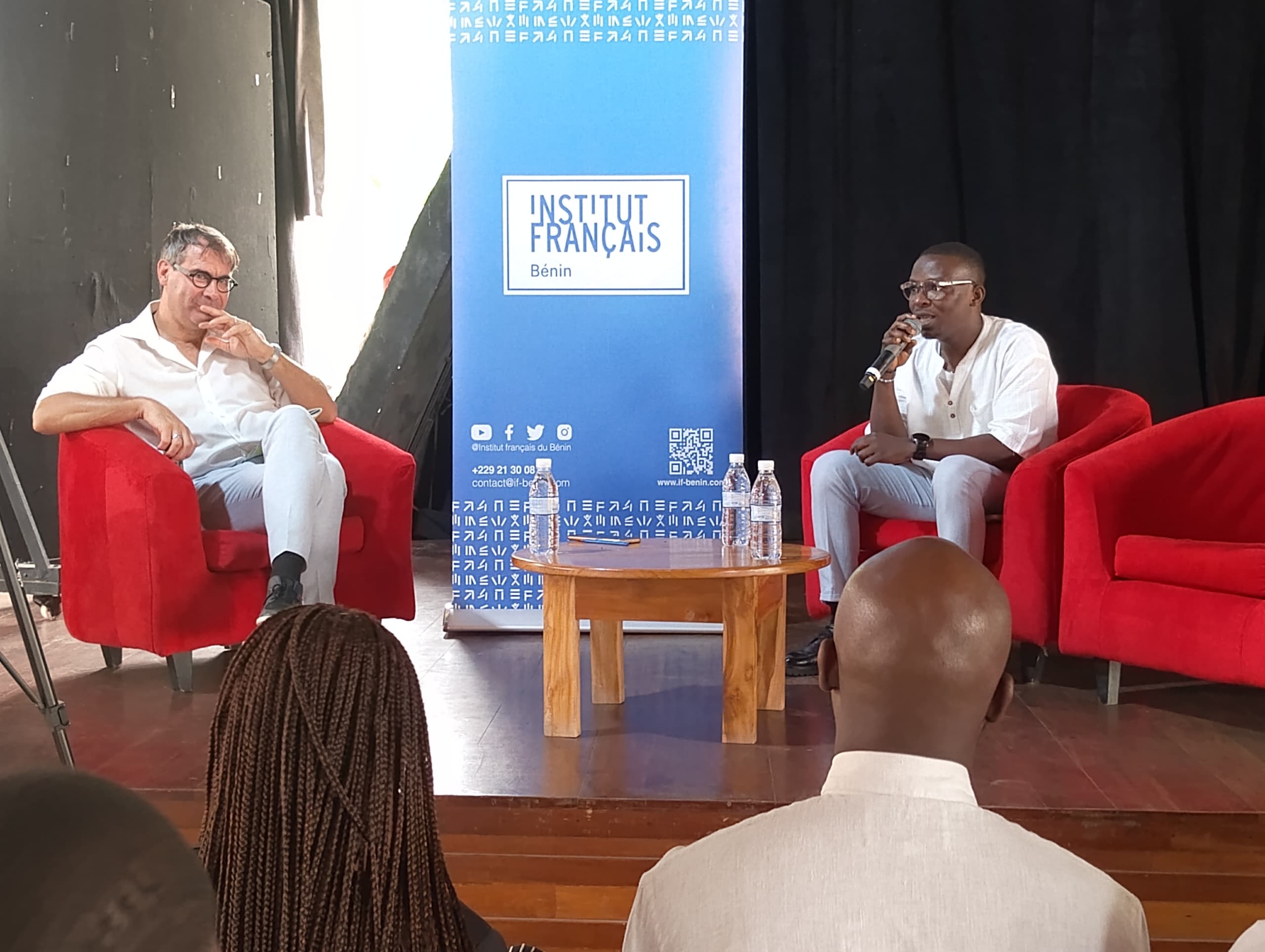Deux hommes discutent sur scène assis dans des fauteuils rouges avec un public et un panneau Institut Français en arrière-plan
