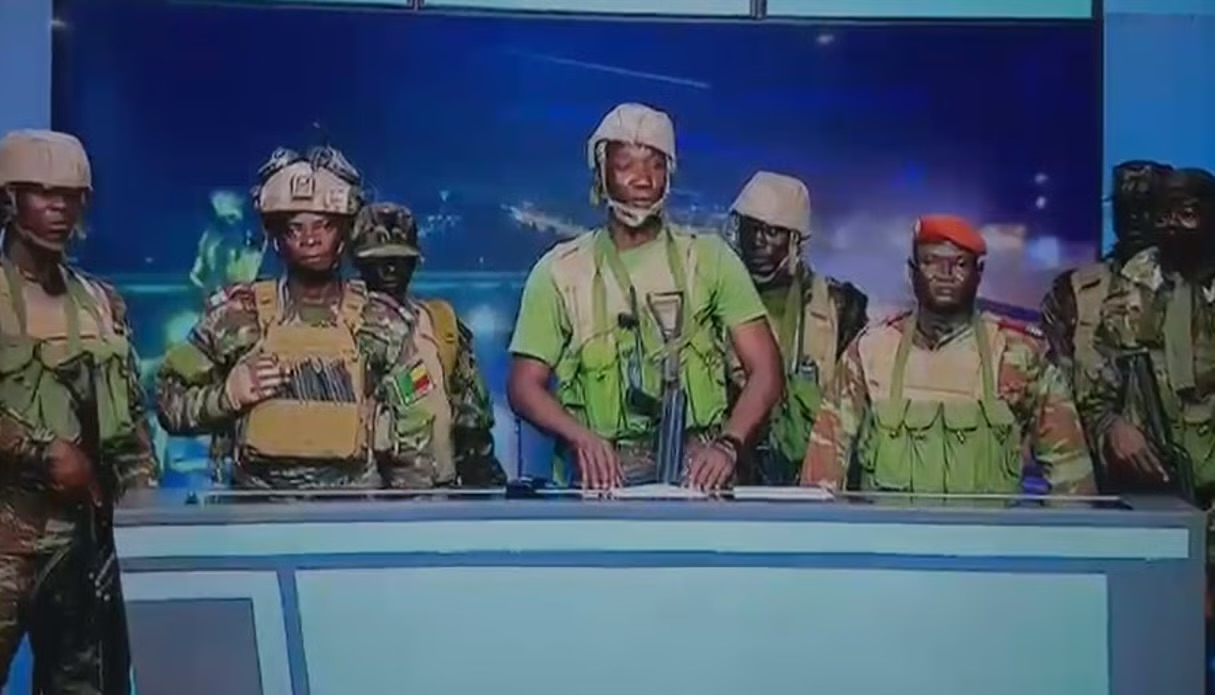 Des hommes en tenue militaire et casque se tiennent derrière un pupitre dans un studio télévisé au fond bleu