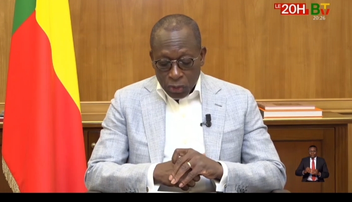 Un homme en veste claire lit un discours assis devant un drapeau tricolore rouge jaune vert avec un interprète en langue des signes en bas à droite