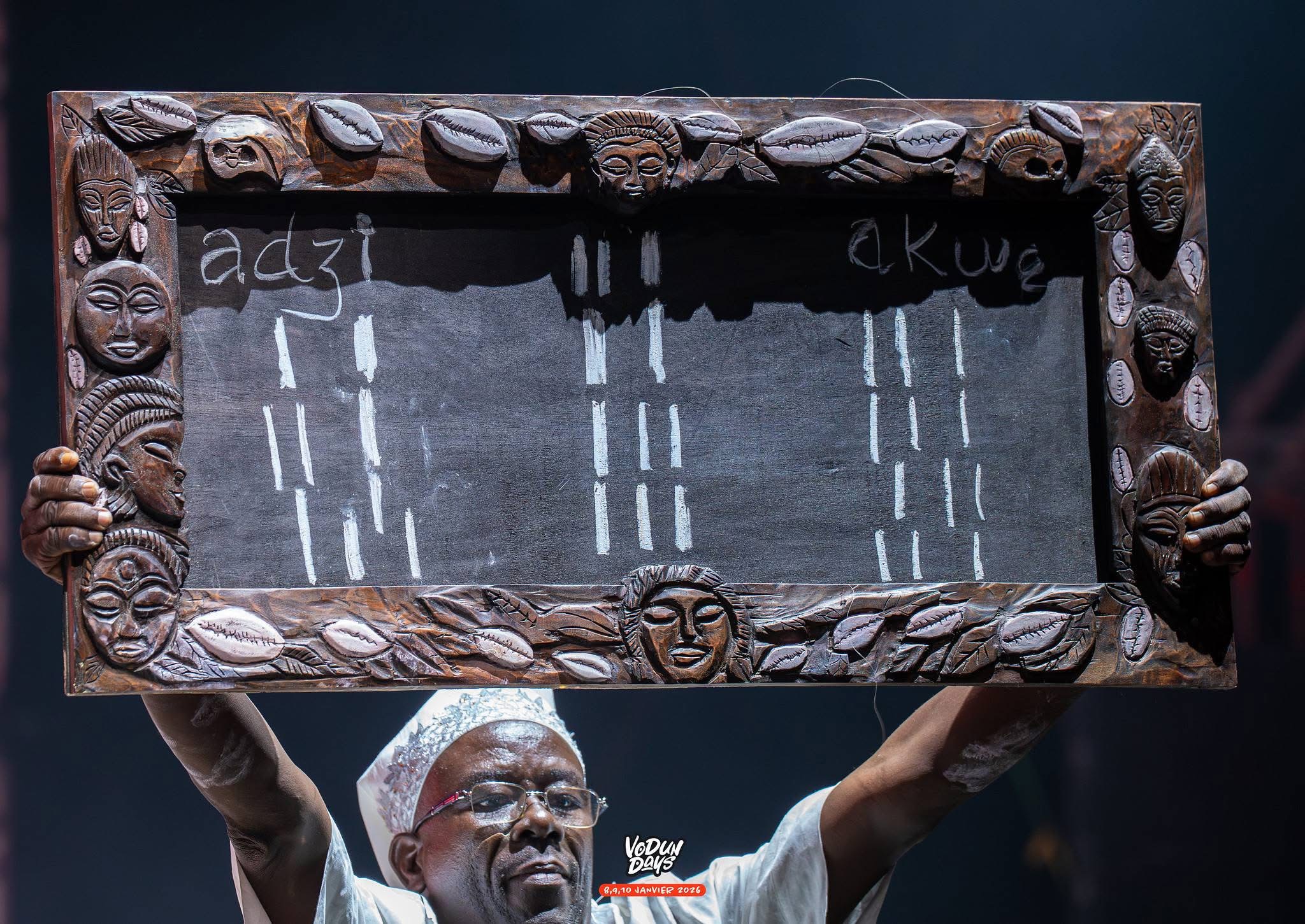 Un homme tient en hauteur un tableau noir encadré de sculptures représentant des visages et des motifs traditionnels avec des marques au craie blanche