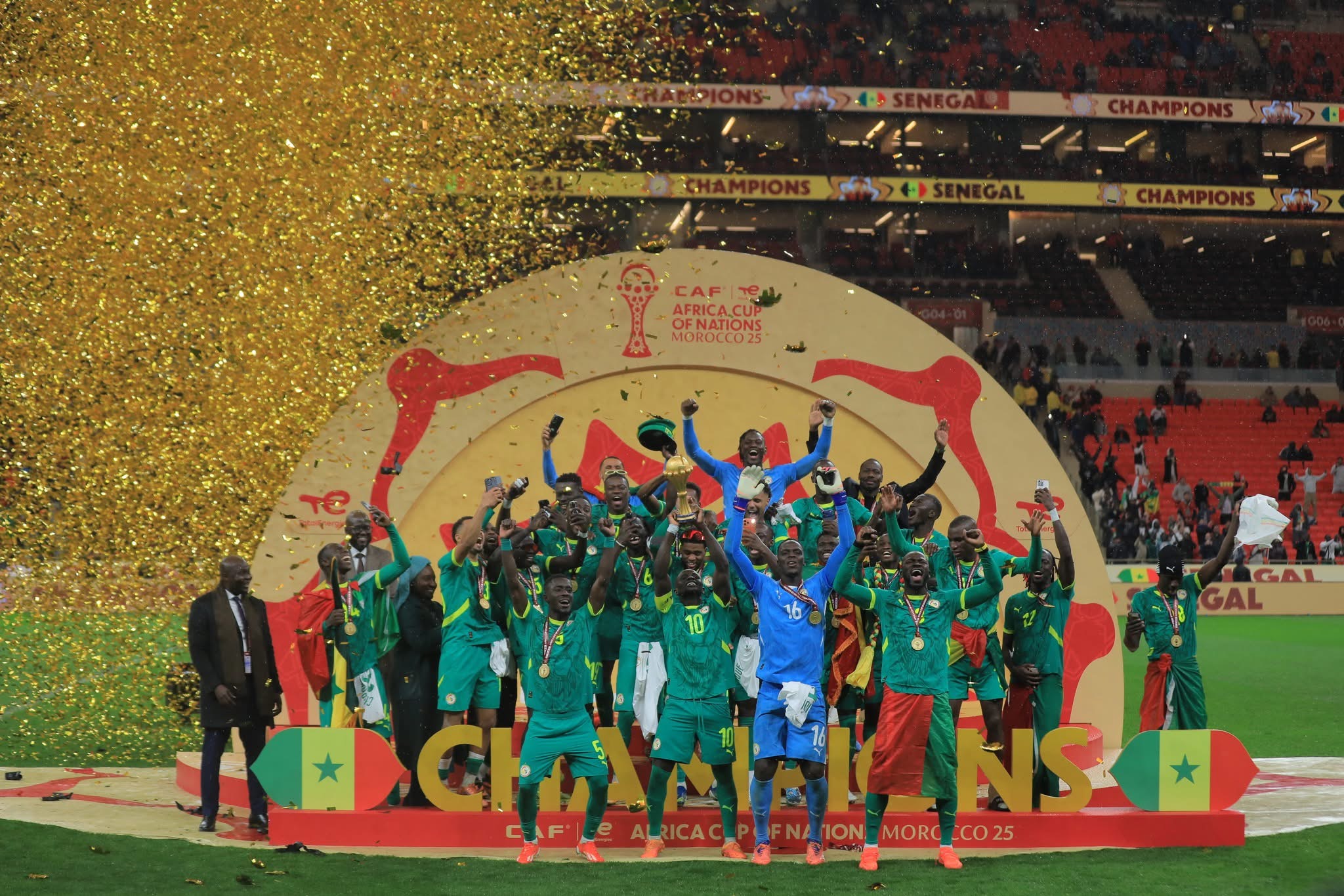 Équipe de football en vert célébrant un trophée sur un podium avec confettis dorés dans un stade rempli de spectateurs