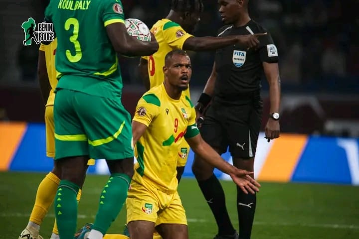 Un joueur en jaune agenouillé et gesticulant face à deux joueurs en vert et un arbitre sur un terrain de football en pleine action