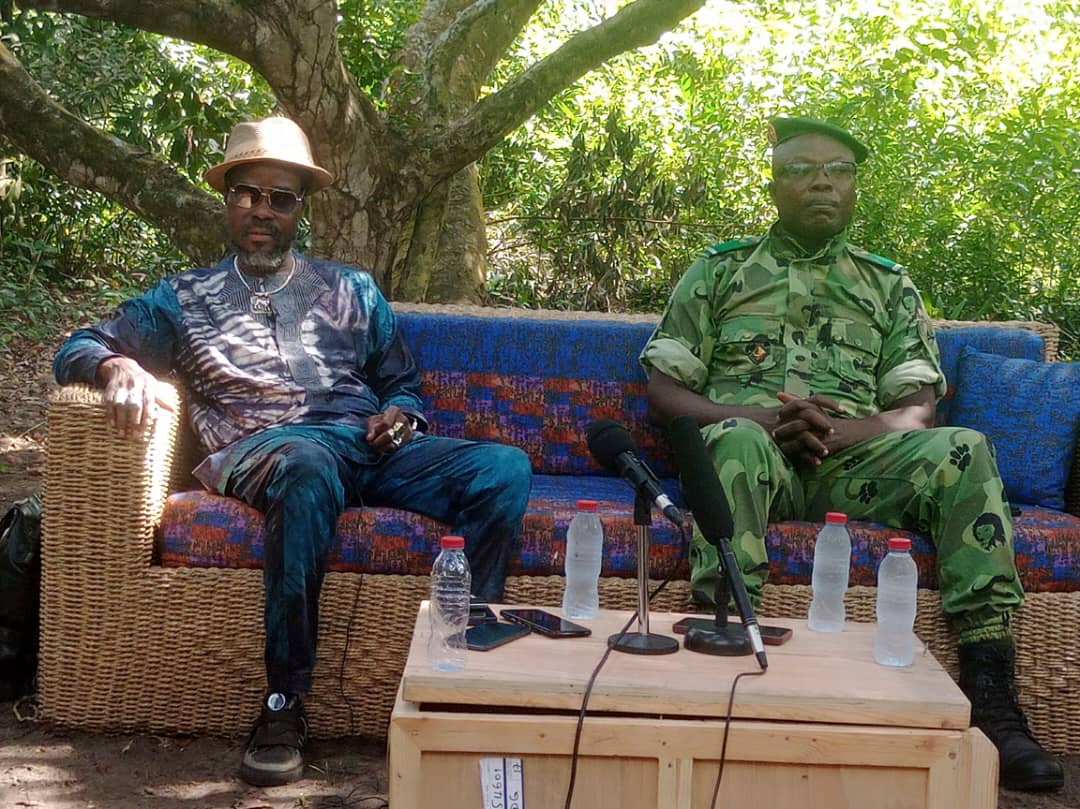 Deux hommes assis sur un canapé en osier sous un arbre, avec plusieurs bouteilles d'eau et des micros sur une table basse devant eux
