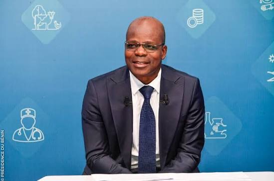 Homme en costume sombre et cravate assis devant un fond bleu avec des icônes blanches liées à la santé et l'économie