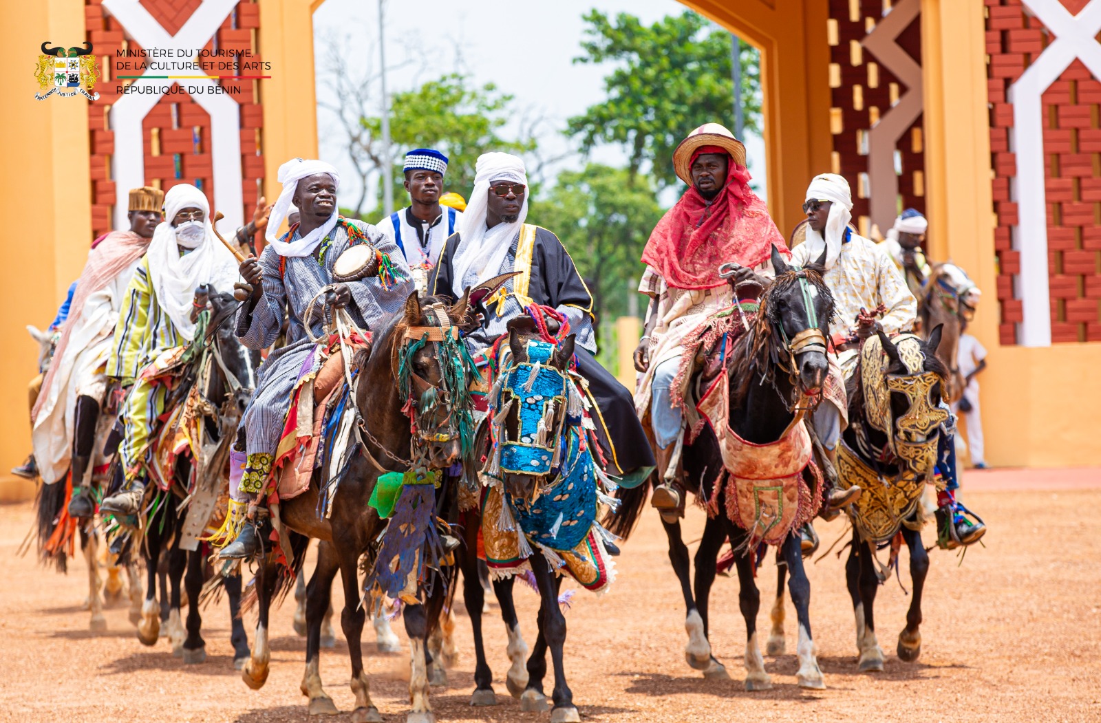 Un groupe d’hommes en habits traditionnels colorés montent des chevaux richement décorés au sol rouge devant un bâtiment aux murs ocres