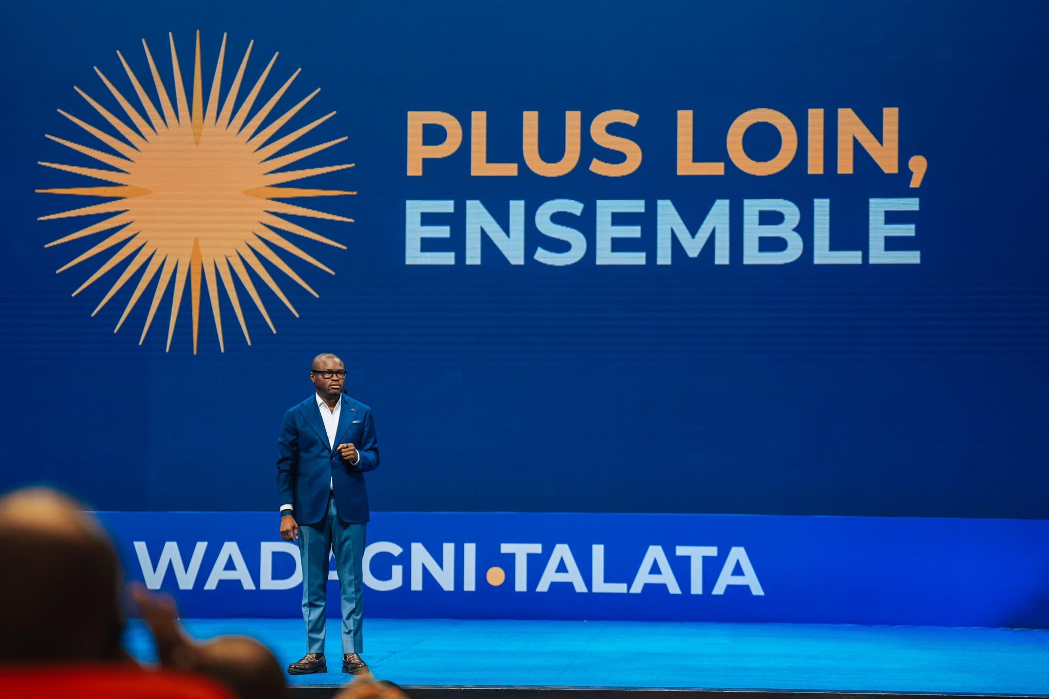 Un homme en costume debout sur scène devant un grand écran bleu avec le texte « Plus loin, ensemble