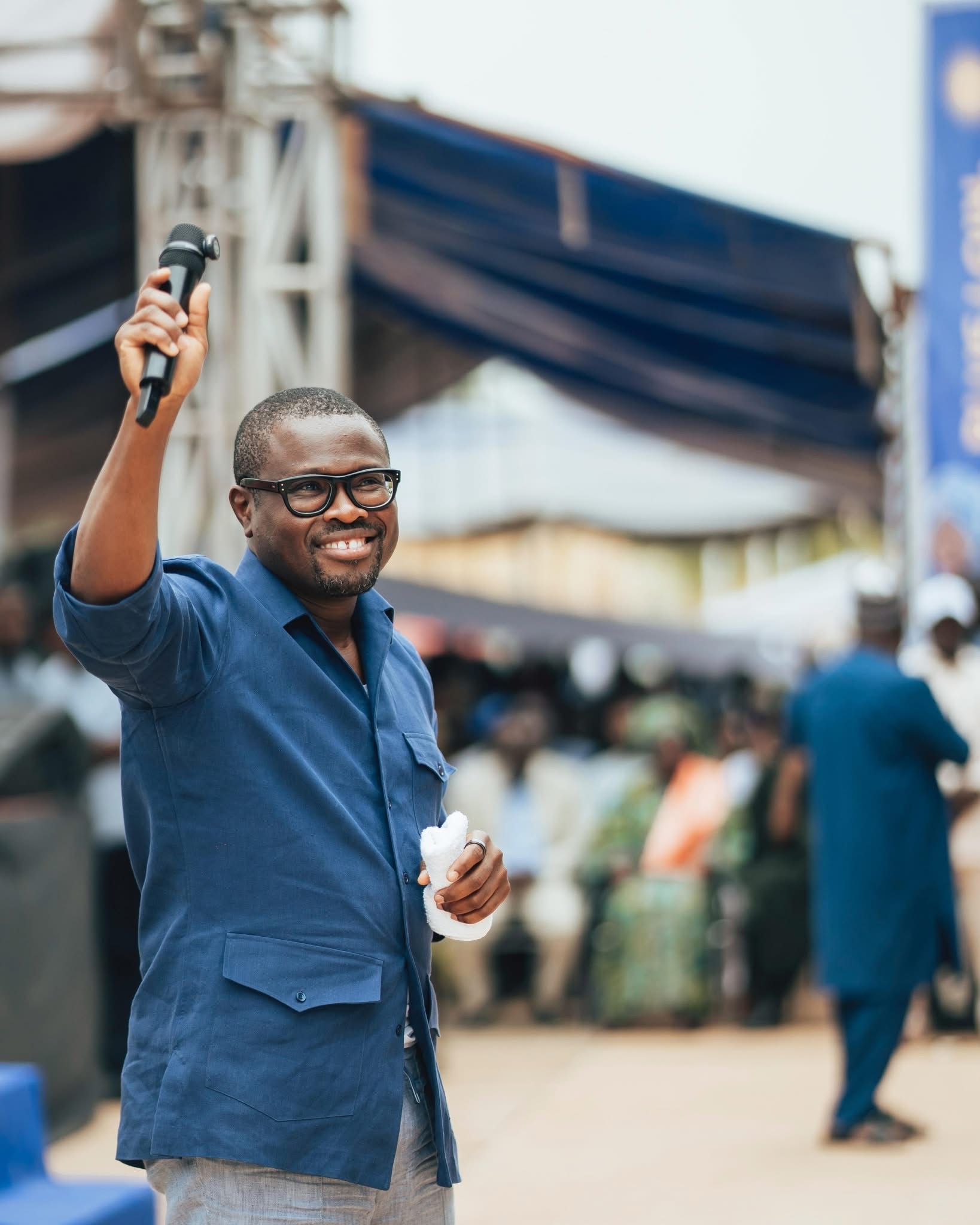Homme souriant en chemise bleue lunettes noires tenant un micro et un tissu blanc lors d'un rassemblement en plein air
