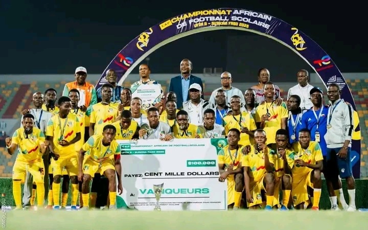 Équipe de football scolaire posant avec un grand chèque et un trophée sous un arc aux couleurs de la compétition africaine