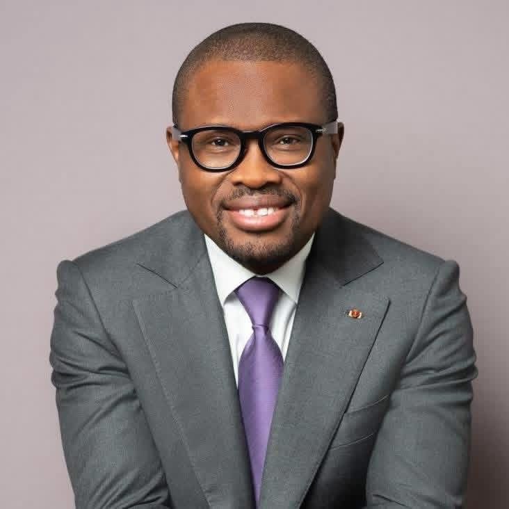 Homme souriant portant un costume gris, une cravate violette et des lunettes noires sur fond uni gris