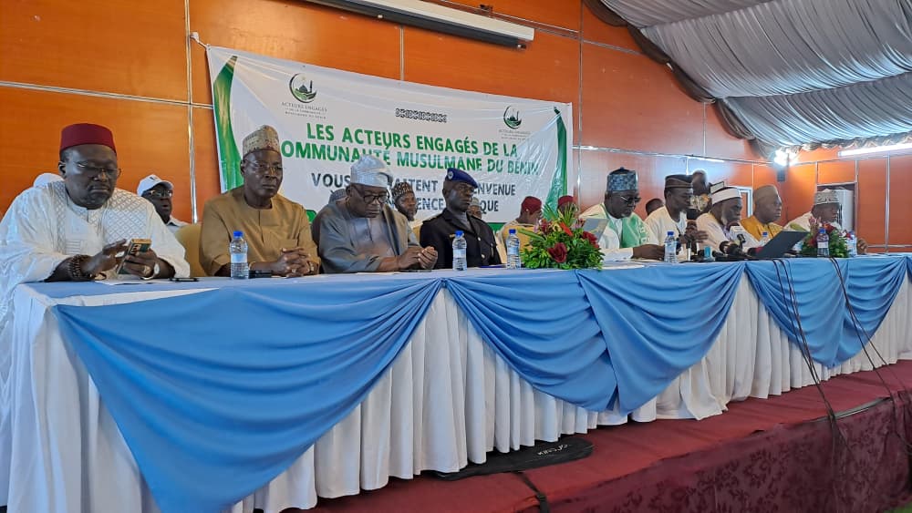 Huit hommes vêtus de tenues traditionnelles sont assis derrière une table recouverte de nappes blanche et bleue sous une tente avec une banderole en arrière-pla