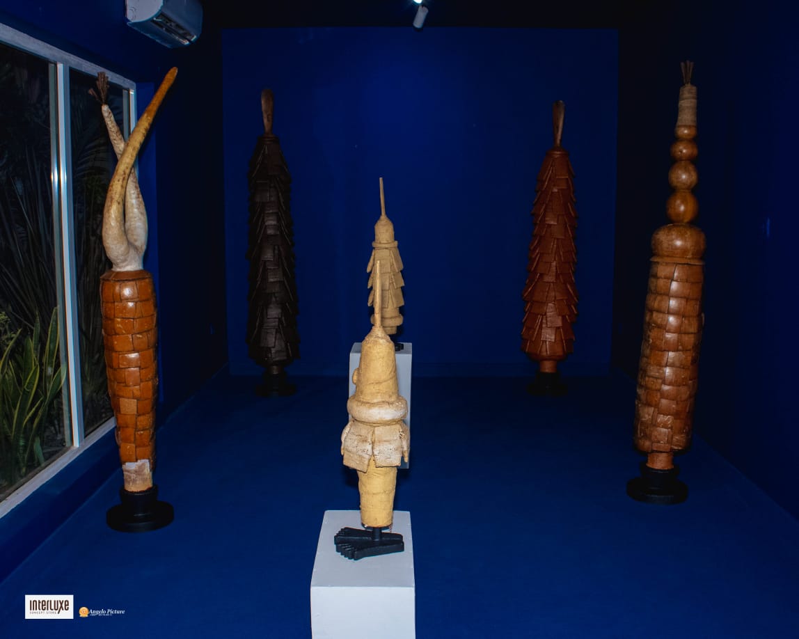 Cinq sculptures hautes en bois placées dans une pièce aux murs et au sol bleus avec une grande vitre à gauche