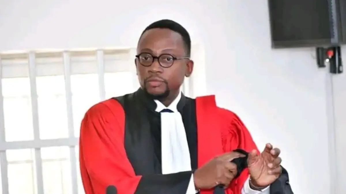 Homme en robe d’avocat rouge et noire ajustant ses manches dans une salle claire avec fenêtre et écran mural