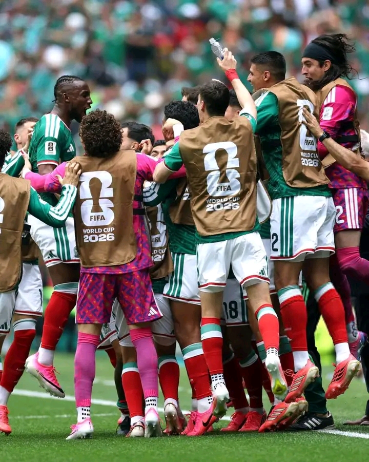 Des joueurs en tenues vertes et rouges célèbrent un but lors d’un match de football sur un terrain gazonné devant un public flou