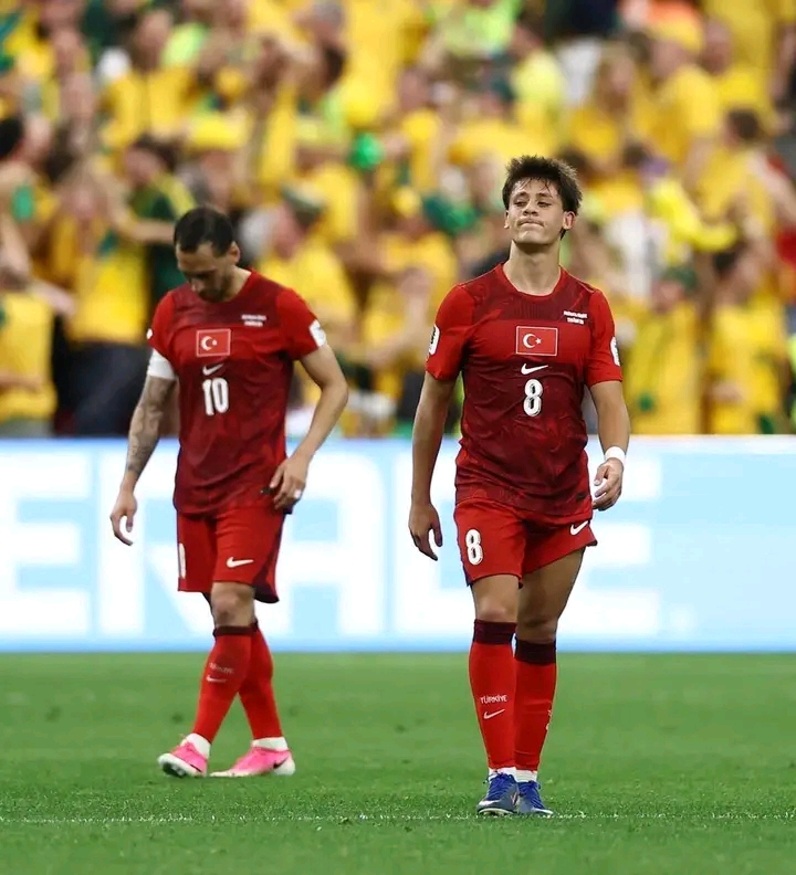 Deux joueurs de football en tenue rouge avec un drapeau sur le maillot avancent avec une expression déçue sur un terrain vert devant des spectateurs en jaune