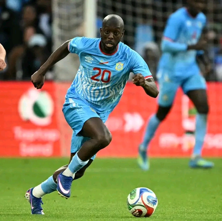 Un joueur en maillot bleu clair dribble un ballon de football sur un terrain en gazon devant une autre personne floue en arrière-plan