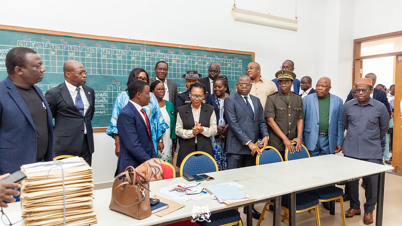 Un groupe de personnes en tenue formelle se tient debout devant un tableau noir dans une salle de classe avec des piles de documents sur une table.