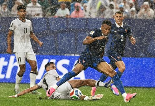 Un joueur en maillot foncé dribble sous la pluie en évitant un adversaire au sol en maillot blanc pendant un match de football