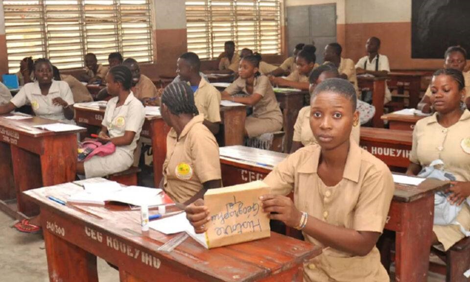 Des élèves en uniforme beige assis dans une salle de classe avec des cahiers et un pupitre en bois marqués