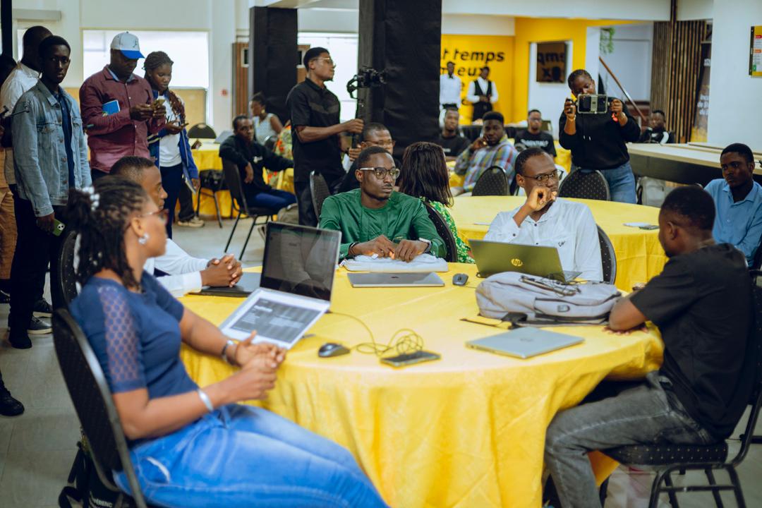 Plusieurs personnes assises autour d’une table jaune discutent et utilisent des ordinateurs portables dans une salle animée