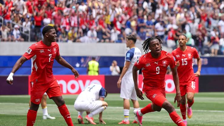 Des joueurs en maillots rouges célèbrent un but lors d’un match de football devant un public dense et des adversaires en blanc désabusés