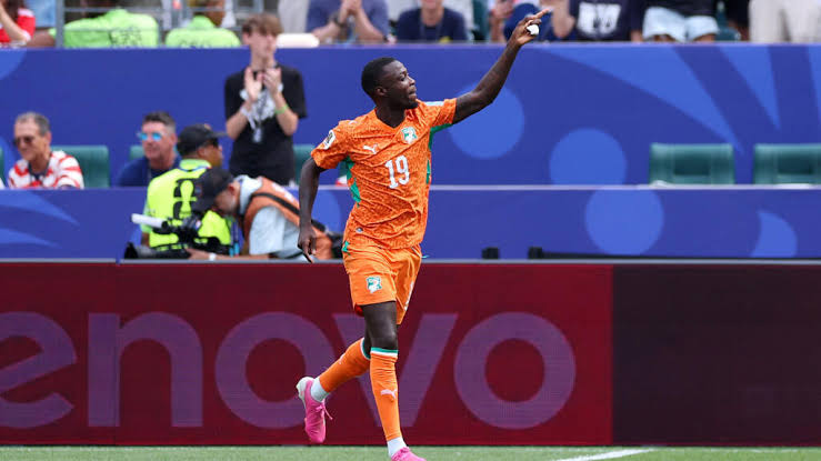 Joueur de football en maillot orange levant le bras lors d'une célébration sur le terrain devant des spectateurs applaudissant